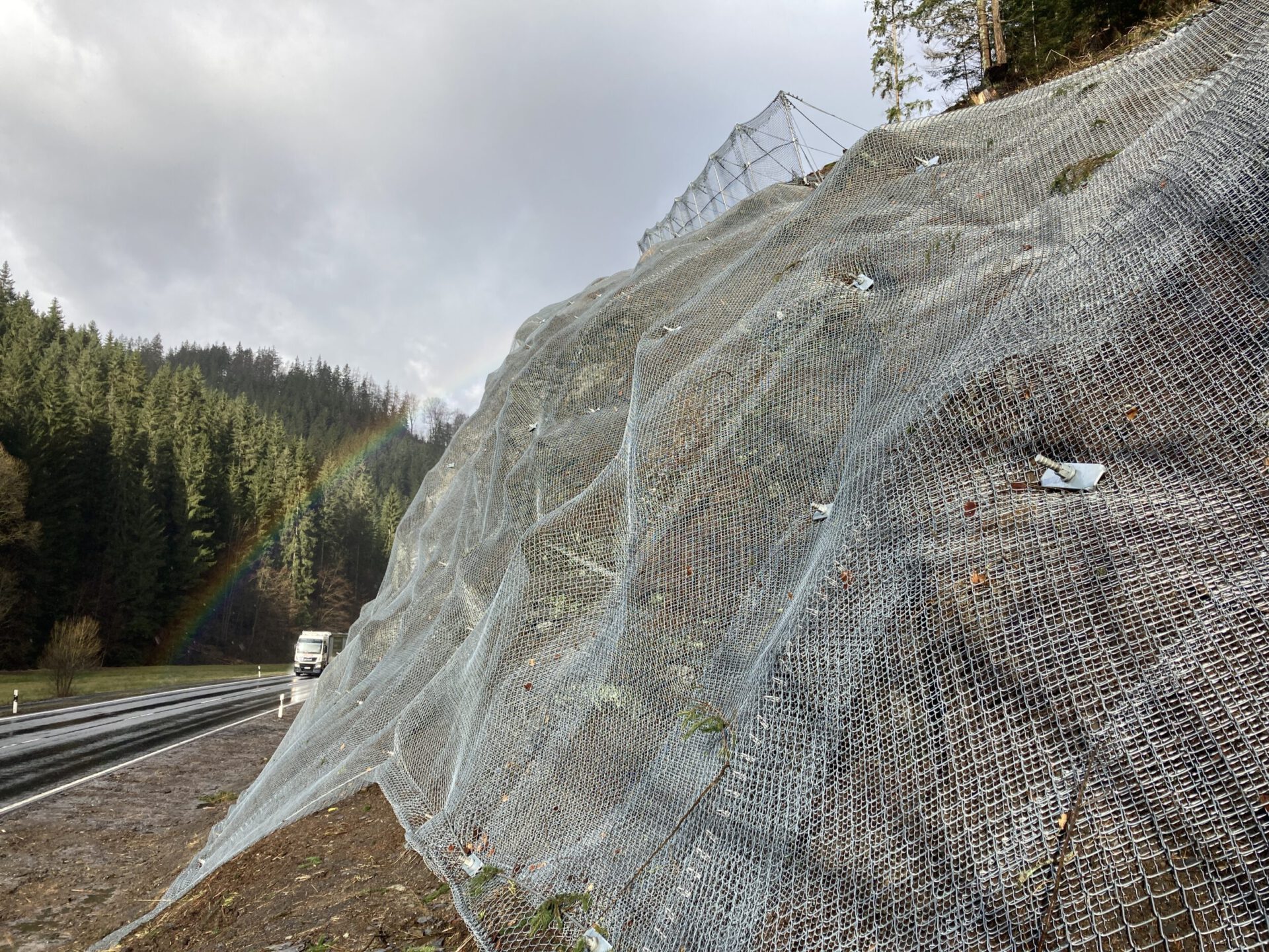 Das Bild zeigt eine steile Straßenböschung, die vollständig mit einem engmaschigen Drahtnetz gesichert ist. Das Netz liegt in mehreren überlappenden Schichten über der Böschung und wird durch Befestigungspunkte stabilisiert. Links verläuft eine Straße mit einem Lkw, dahinter erstreckt sich ein dichter Wald, und im Hintergrund ist ein Regenbogen vor einem bewölkten Himmel zu sehen.