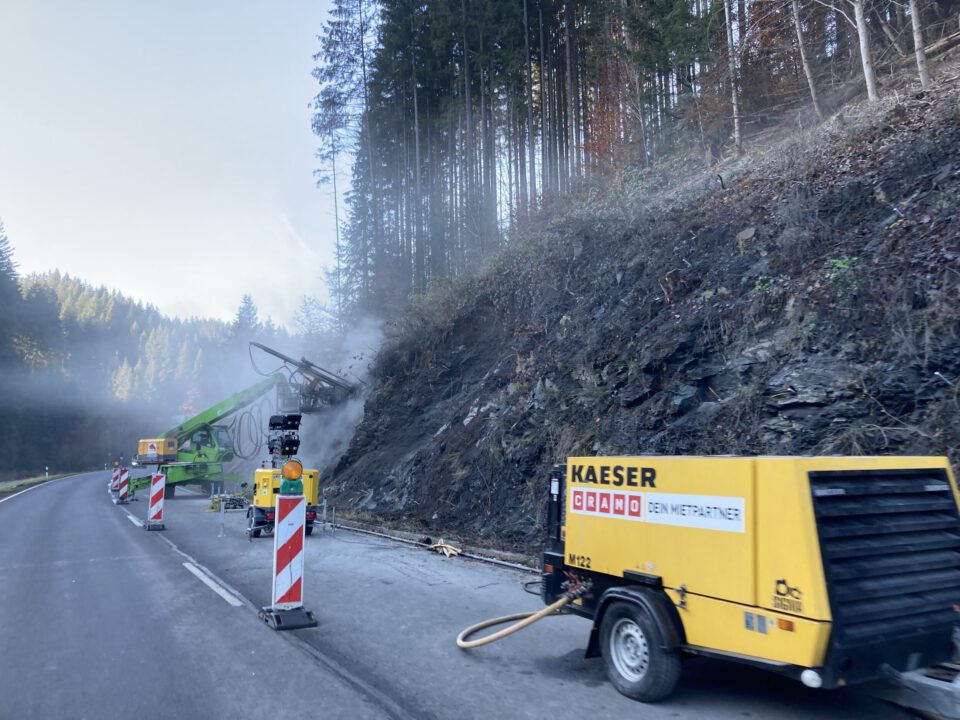 Das Bild zeigt eine Baustelle an einer kurvigen Landstraße in einem bewaldeten Tal. Rechts im Vordergrund steht ein gelber mobiler Kompressor, von dem ein Schlauch zur Bohrstelle führt. Die Straße ist durch rot-weiße Leitbaken und Warntechnik abgesichert. Links daneben befindet sich ein grünes Bohrgerät mit langem Ausleger, das direkt in die dunkle, felsige Böschung bohrt. Aus der Bohrstelle steigt Staub oder Dampf auf. Vor der Maschine steht ein gelbes Warn- und Steuergerät mit Blinklicht.