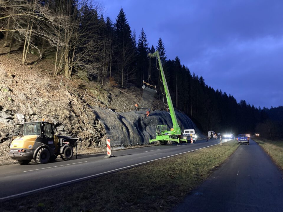 Das Bild zeigt eine nächtliche Baustellenszene an einer Landstraße entlang eines steilen bewaldeten Hangs. Rechts verläuft die zweispurige Straße, auf der mehrere Fahrzeuge mit eingeschaltetem Licht stehen oder fahren. Links die Gegenfahrbahn mit Baustellenabsicherung durch rot-weiße Leitbaken. Im Zentrum steht ein großer grüner Teleskopkran, dessen Ausleger schräg nach oben zum Hang reicht. Am Kran hängt eine Netz- bzw. Mattenkonstruktion, die am Fels befestigt wird. Auf dem Hang sind mehrere Arbeiter in orangefarbener Schutzkleidung zu erkennen, die an der Sicherung der Felswand arbeiten.