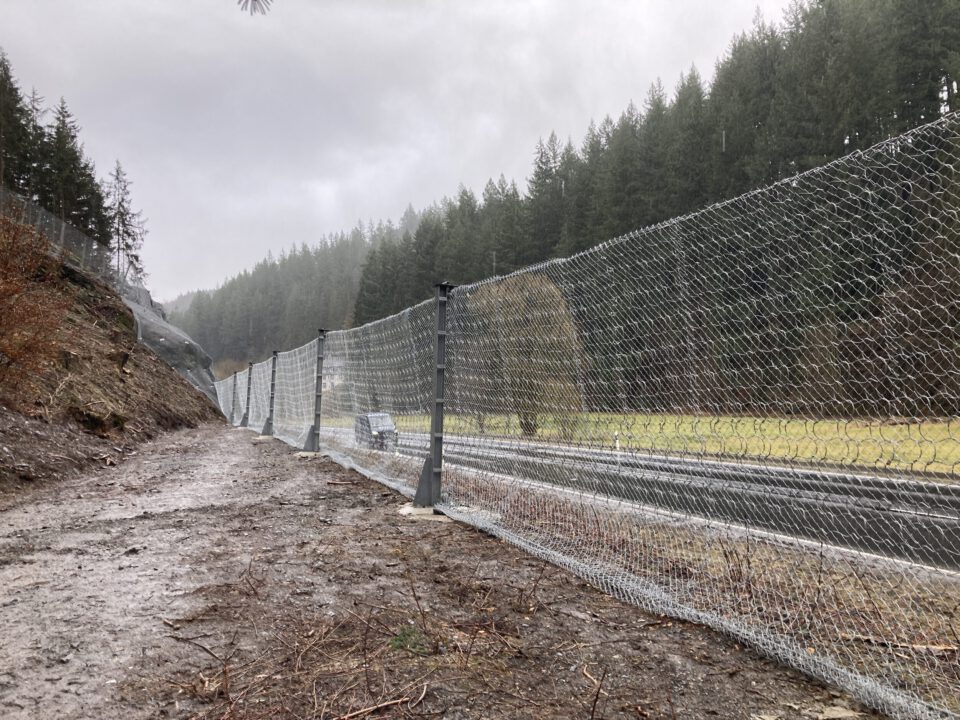 Das Bild zeigt einen Steinschlagschutzzaun, der entlang einer Straße verläuft und aus einem stabilen Drahtgeflecht besteht, das an mehreren Metallpfosten befestigt ist. Links befindet sich eine Böschung mit Erde und Sträuchern, während rechts die Fahrbahn sichtbar ist, die von einem Fahrzeug genutzt wird. Im Hintergrund erstreckt sich ein dichter Wald aus Nadelbäumen unter einem grauen, regnerischen Himmel, was auf eine feuchte Witterung hinweist.