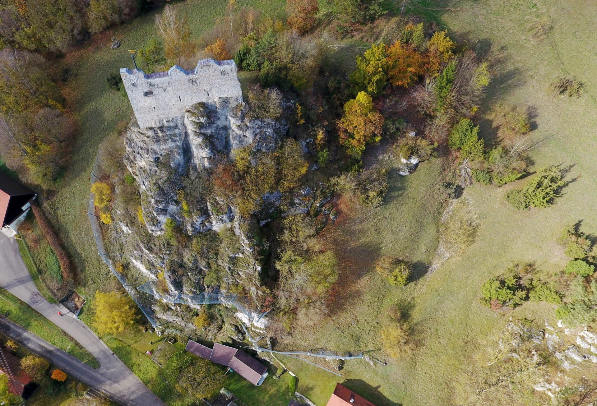 Das Bild zeigt eine große Felsformation, auf deren Spitze die Ruine einer Burg steht. Der Felsen ist von Bäumen und Sträuchern umgeben, die herbstlich gefärbt sind, mit Grün-, Gelb- und Orangetönen. Am unteren Rand des Hangs verläuft ein Steinschlagschutzzaun, und in der Umgebung sind mehrere Gebäude sowie eine Straße sichtbar, die sich durch die Landschaft schlängelt.