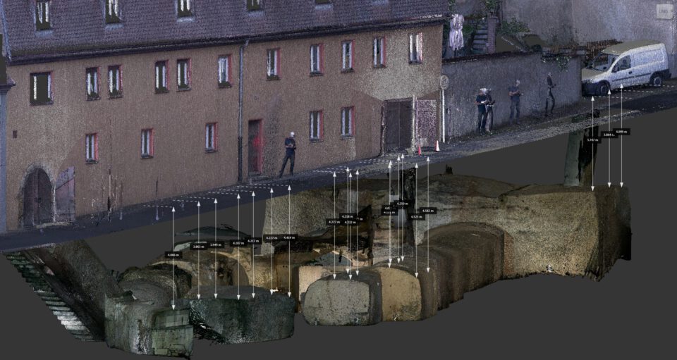 Das Bild zeigt eine digitale 3D-Darstellung, die die Front eines Gebäudes und das darunterliegende Kellersystem kombiniert. Oben ist die Außenansicht eines langgestreckten Gebäudes mit mehreren Fenstern, Türen und einem Fahrzeug auf der Straße zu sehen. Darunter sind gewölbte Kellerstrukturen mit detaillierten Maßangaben dargestellt, die die räumliche Lage und Tiefe der unterirdischen Räume im Verhältnis zur Straßenoberfläche verdeutlichen.