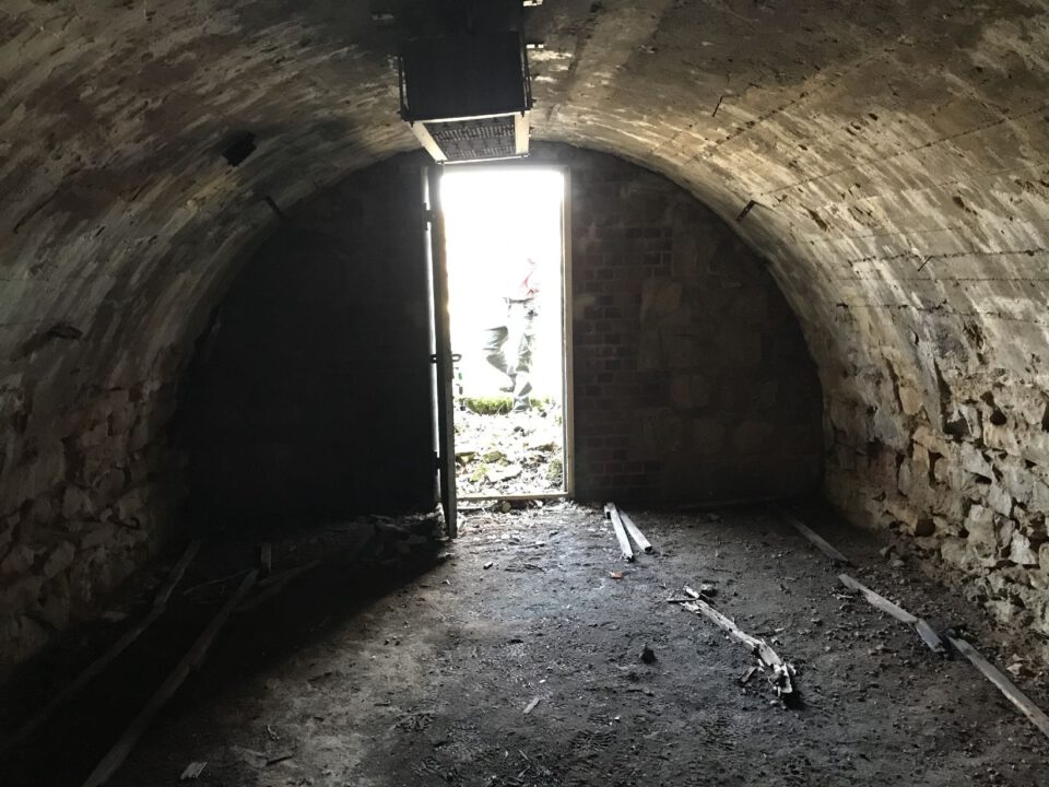 Das Bild zeigt das Innere eines gewölbten Kellerraums mit einer groben Stein- und Betonwand. Der Boden ist uneben und mit Schmutz sowie einigen losen Holz- und Metallteilen bedeckt. Am Ende des Raumes befindet sich eine geöffnete Tür, durch die helles Tageslicht einfällt und den Ausgang nach draußen sichtbar macht.