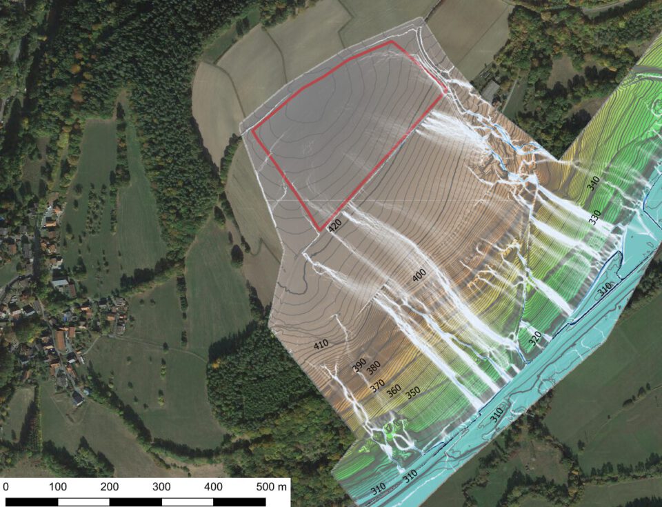 Das Bild zeigt eine Luftaufnahme einer landwirtschaftlich geprägten Landschaft mit Feldern, Waldflächen und einem kleinen Dorf am linken Rand. Über die Aufnahme ist ein farbiges Modell gelegt, das Höhenlinien und Abflusswege darstellt. Die Höhenlinien sind mit Zahlen markiert, die die Höhenmeter angeben, und die farbigen Bereiche reichen von Braun über Grün bis Blau, was unterschiedliche Höhenstufen und Wasserfließrichtungen verdeutlicht.