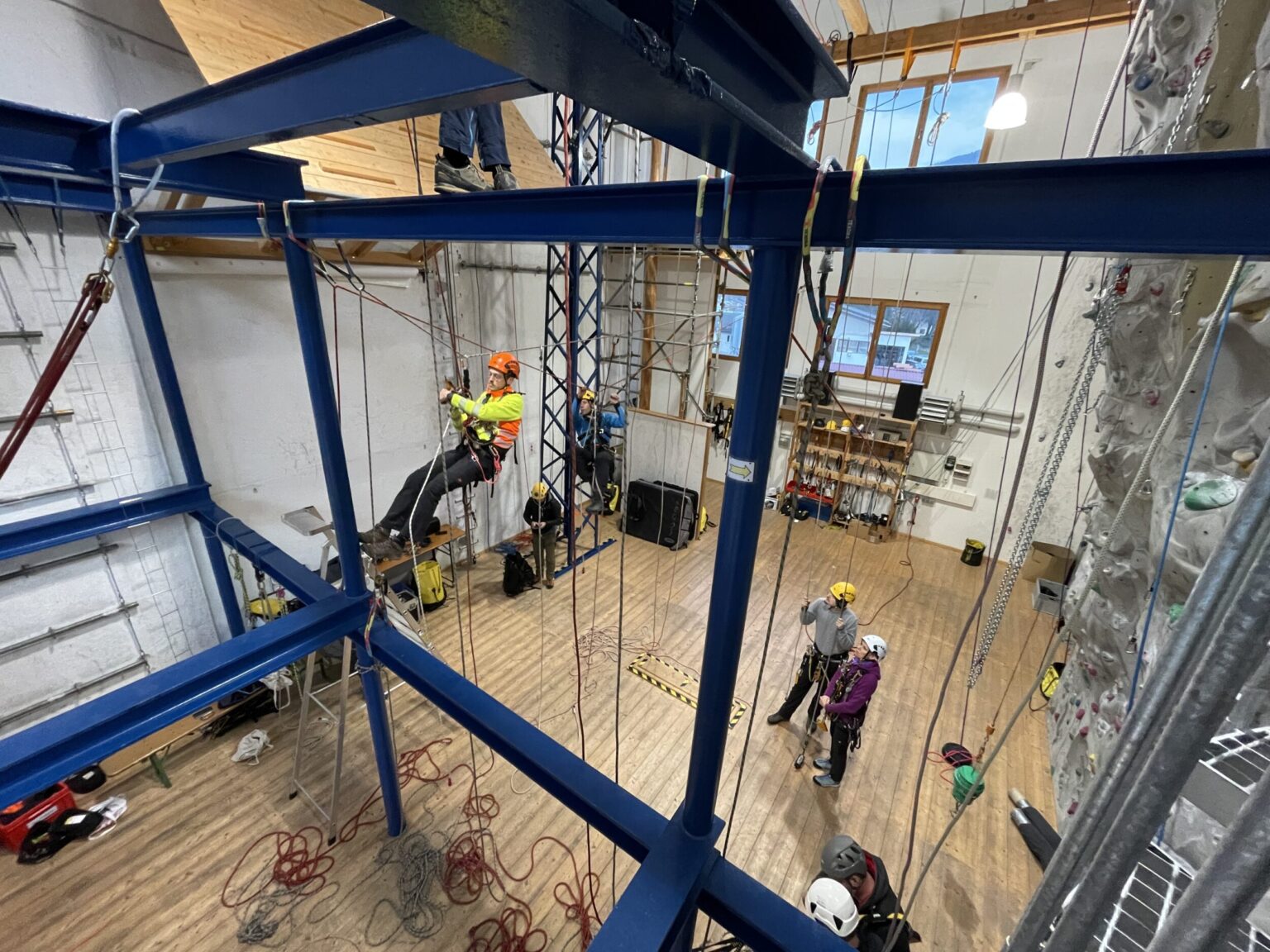 Das Bild zeigt eine große Halle mit einem stabilen blauen Stahlgerüst, an dem mehrere Seile befestigt sind. Eine Person hängt gesichert in einem Klettergurt am Gerüst, während andere Personen mit Helmen und Ausrüstung am Boden stehen und offenbar eine Übung oder Schulung beobachten. Im Hintergrund sind eine Kletterwand, Holzregale mit Ausrüstung und große Fenster zu sehen, die für Tageslicht sorgen.
