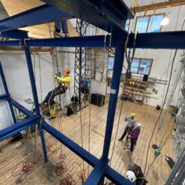 Das Bild zeigt eine große Halle mit einem stabilen blauen Stahlgerüst, an dem mehrere Seile befestigt sind. Eine Person hängt gesichert in einem Klettergurt am Gerüst, während andere Personen mit Helmen und Ausrüstung am Boden stehen und offenbar eine Übung oder Schulung beobachten. Im Hintergrund sind eine Kletterwand, Holzregale mit Ausrüstung und große Fenster zu sehen, die für Tageslicht sorgen.