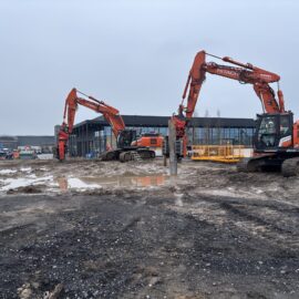 Auf dem Bild ist eine Baustelle mit zwei großen orangefarbenen Baggern zu sehen, die auf schlammigem Boden stehen. Im Hintergrund befindet sich ein modernes, langgestrecktes Gebäude mit großen Glasfronten, daneben sind Absperrungen und weitere Baustellenelemente erkennbar. Der Himmel ist grau und bewölkt, was auf trübes Wetter hinweist.