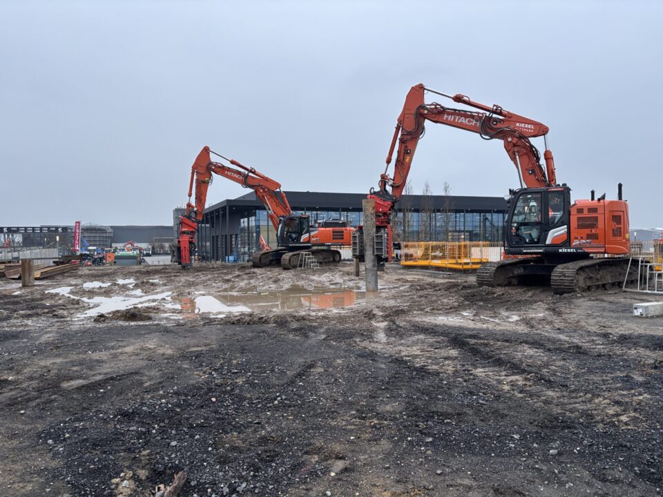 Auf dem Bild ist eine Baustelle mit zwei großen orangefarbenen Baggern zu sehen, die auf schlammigem Boden stehen. Im Hintergrund befindet sich ein modernes, langgestrecktes Gebäude mit großen Glasfronten, daneben sind Absperrungen und weitere Baustellenelemente erkennbar. Der Himmel ist grau und bewölkt, was auf trübes Wetter hinweist.