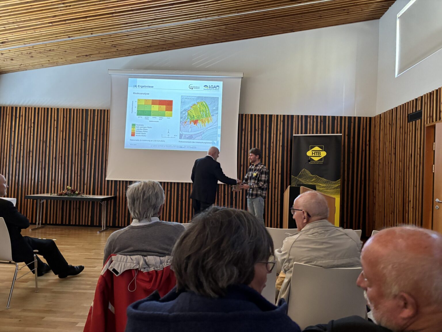 Das Bild zeigt einen Vortrag in einem modernen Konferenzraum mit Holzverkleidung an den Wänden und einer schrägen Holzdecke. Im Vordergrund sitzen mehrere Personen auf weißen Stühlen und hören zu. Im Hintergrund ist eine große Leinwand mit einer Präsentation zu sehen, die farbige Diagramme und Karten enthält, sowie ein Banner mit einem gelben Logo. Zwei Personen stehen vorne und scheinen sich die Hand zu geben, vermutlich im Rahmen einer Begrüßung oder Übergabe.