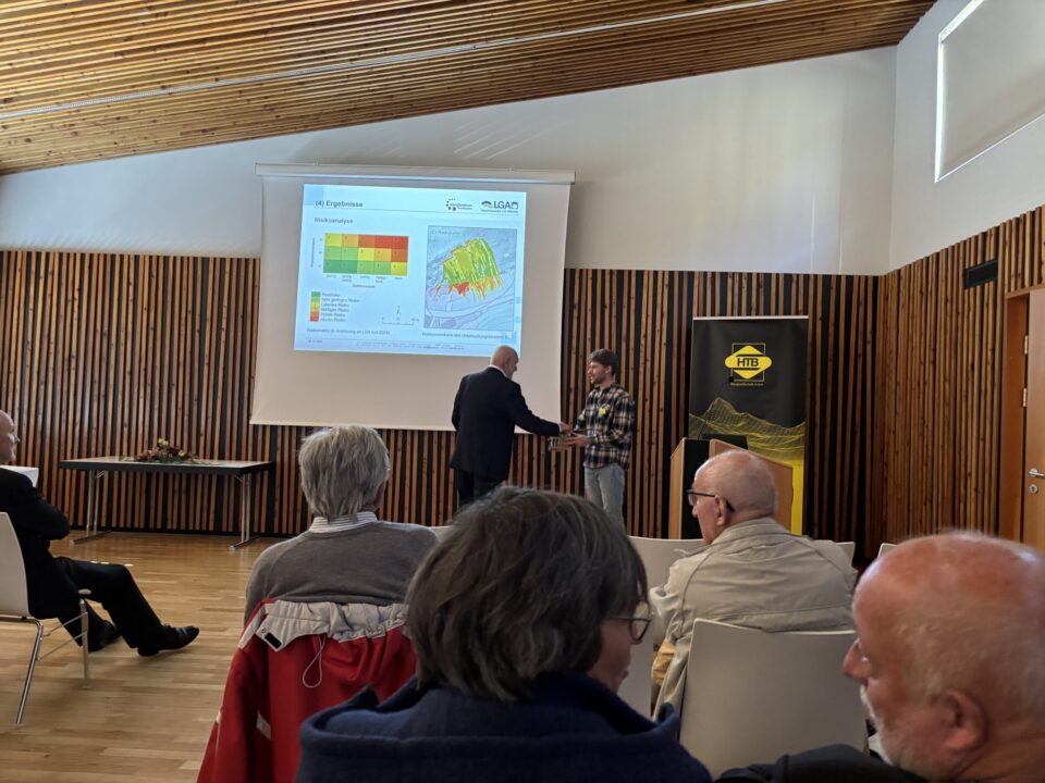 Das Bild zeigt einen Vortrag in einem modernen Konferenzraum mit Holzverkleidung an den Wänden und einer schrägen Holzdecke. Im Vordergrund sitzen mehrere Personen auf weißen Stühlen und hören zu. Im Hintergrund ist eine große Leinwand mit einer Präsentation zu sehen, die farbige Diagramme und Karten enthält, sowie ein Banner mit einem gelben Logo. Zwei Personen stehen vorne und scheinen sich die Hand zu geben, vermutlich im Rahmen einer Begrüßung oder Übergabe.