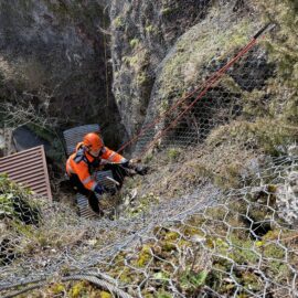 Das Bild zeigt eine steile Felsböschung, die mit einem Drahtnetz zur Sicherung gegen Steinschlag versehen ist. Eine Person in orangefarbener Schutzkleidung und Helm ist mit Seilen gesichert und arbeitet am Hang, offenbar bei der Kontrolle oder Wartung des Netzes. Im Hintergrund sind Felsen, Vegetation und einige Dachflächen von Gebäuden zu erkennen.