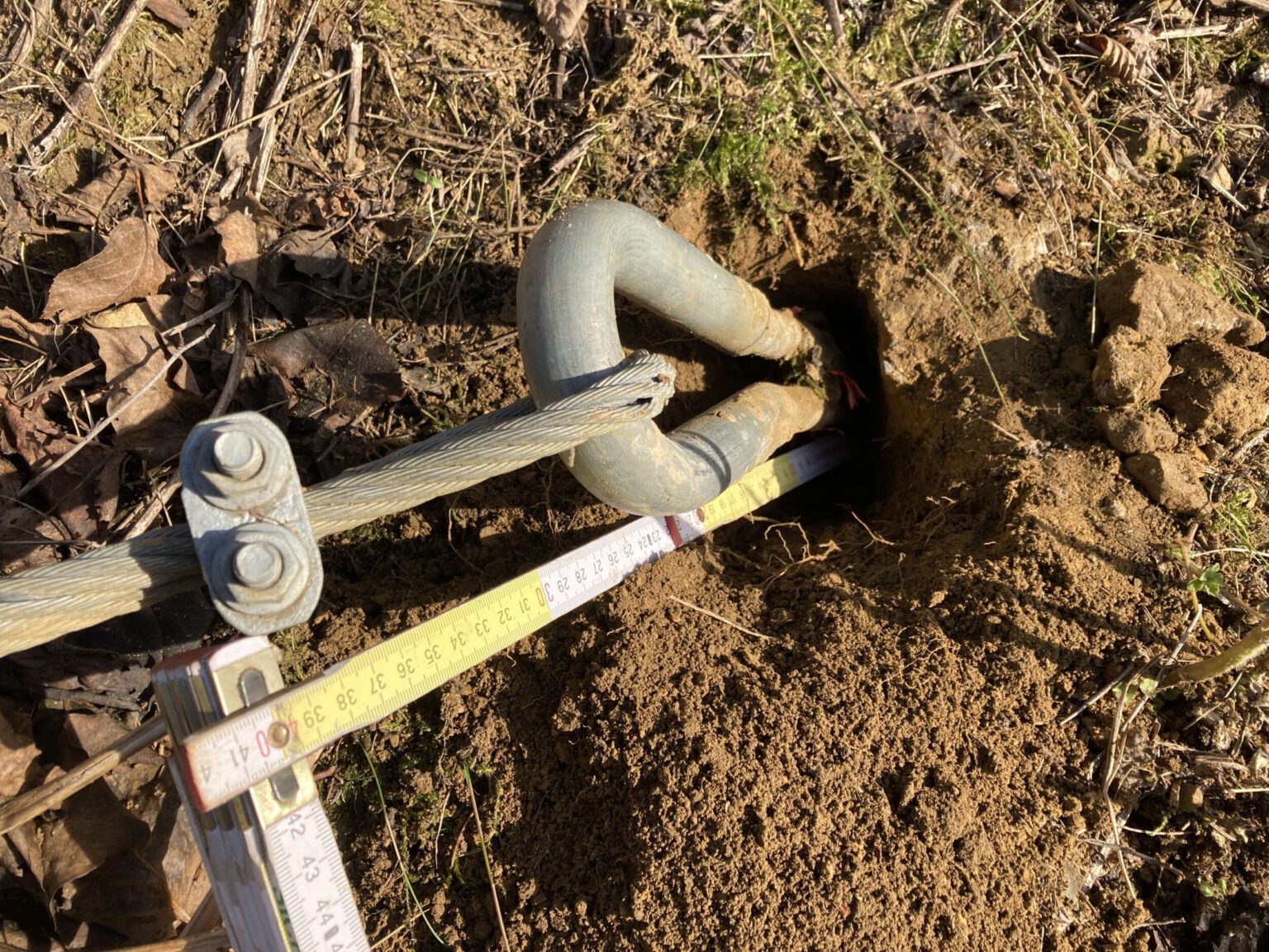 Das Bild zeigt ein teilweise freigelegtes Metallbauteil, das aus einem Erdloch herausragt und mit einem Drahtseil verbunden ist. Das Bauteil ist silberfarben und gebogen, während das Drahtseil mit einer Klemme fixiert ist. Um das Loch herum liegt lockere, sandige Erde mit einigen trockenen Blättern und Gras, und ein Maßband ist über das Loch gelegt, um die Tiefe oder Position zu messen.