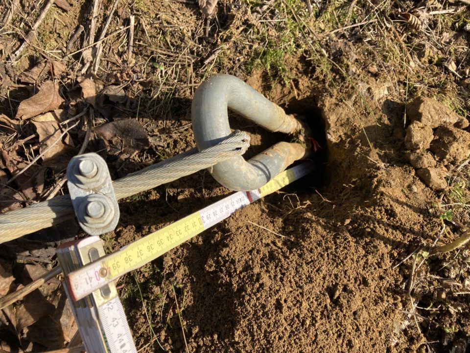 Das Bild zeigt ein teilweise freigelegtes Metallbauteil, das aus einem Erdloch herausragt und mit einem Drahtseil verbunden ist. Das Bauteil ist silberfarben und gebogen, während das Drahtseil mit einer Klemme fixiert ist. Um das Loch herum liegt lockere, sandige Erde mit einigen trockenen Blättern und Gras, und ein Maßband ist über das Loch gelegt, um die Tiefe oder Position zu messen.