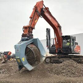 Das Bild zeigt einen großen orangefarbenen Bagger, der auf einem Schotterhaufen arbeitet. Anstelle einer normalen Schaufel ist am Ausleger eine spezielle Siebtrommel angebracht, die Material aufnimmt und durch Drehung sortiert, wobei feiner Schotter nach unten herausfällt. Im Hintergrund sind weitere Baumaschinen, Betonreste und ein großes graues Industriegebäude zu sehen, was auf eine Baustelle oder ein Abbruchgelände hinweist.
