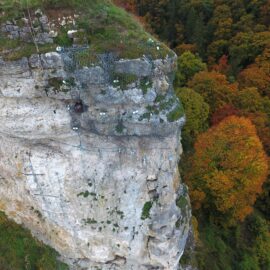 Auf dem Bild ist eine steile Felswand zu sehen, deren oberer Bereich mit Drahtgeflechten und Sicherungselementen versehen ist. Auf dem Felsplateau wächst Gras, und einige Steine liegen lose am Rand. Im Hintergrund erstreckt sich ein dichter Wald mit bunten Herbstfarben in Grün, Gelb und Orange.