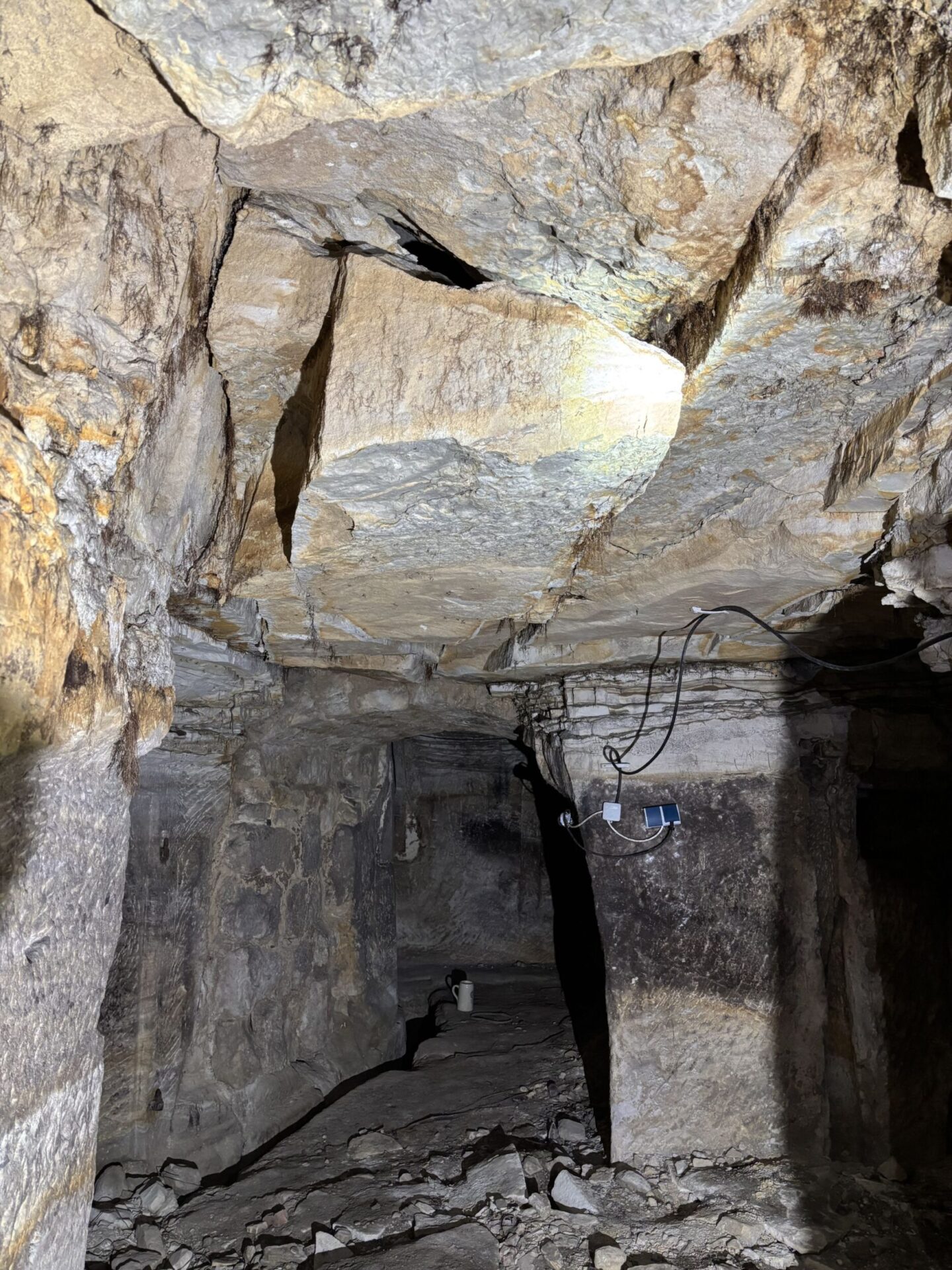 Das Bild zeigt einen unterirdischen Gang mit grob behauenen Felswänden und einer unebenen Decke, in der ein großer Riss sichtbar ist. Mehrere Kabel hängen lose von der Decke, und am Boden liegen zahlreiche Gesteinsbrocken. Die Szene ist mit künstlichem Licht ausgeleuchtet, wodurch die Struktur des brüchigen Gesteins deutlich erkennbar ist.