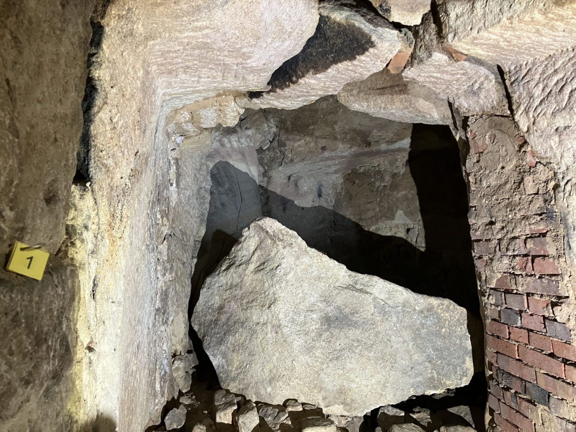 Das Bild zeigt einen unterirdischen Bereich mit grob behauenen Felswänden und einer teilweise gemauerten Wand aus roten Ziegeln. In der Mitte liegt ein großer, abgestürzter Felsblock, umgeben von kleineren Steinen und Geröll. Die Szene ist mit künstlichem Licht ausgeleuchtet, wodurch die Struktur und Bruchkanten des Gesteins deutlich sichtbar sind.