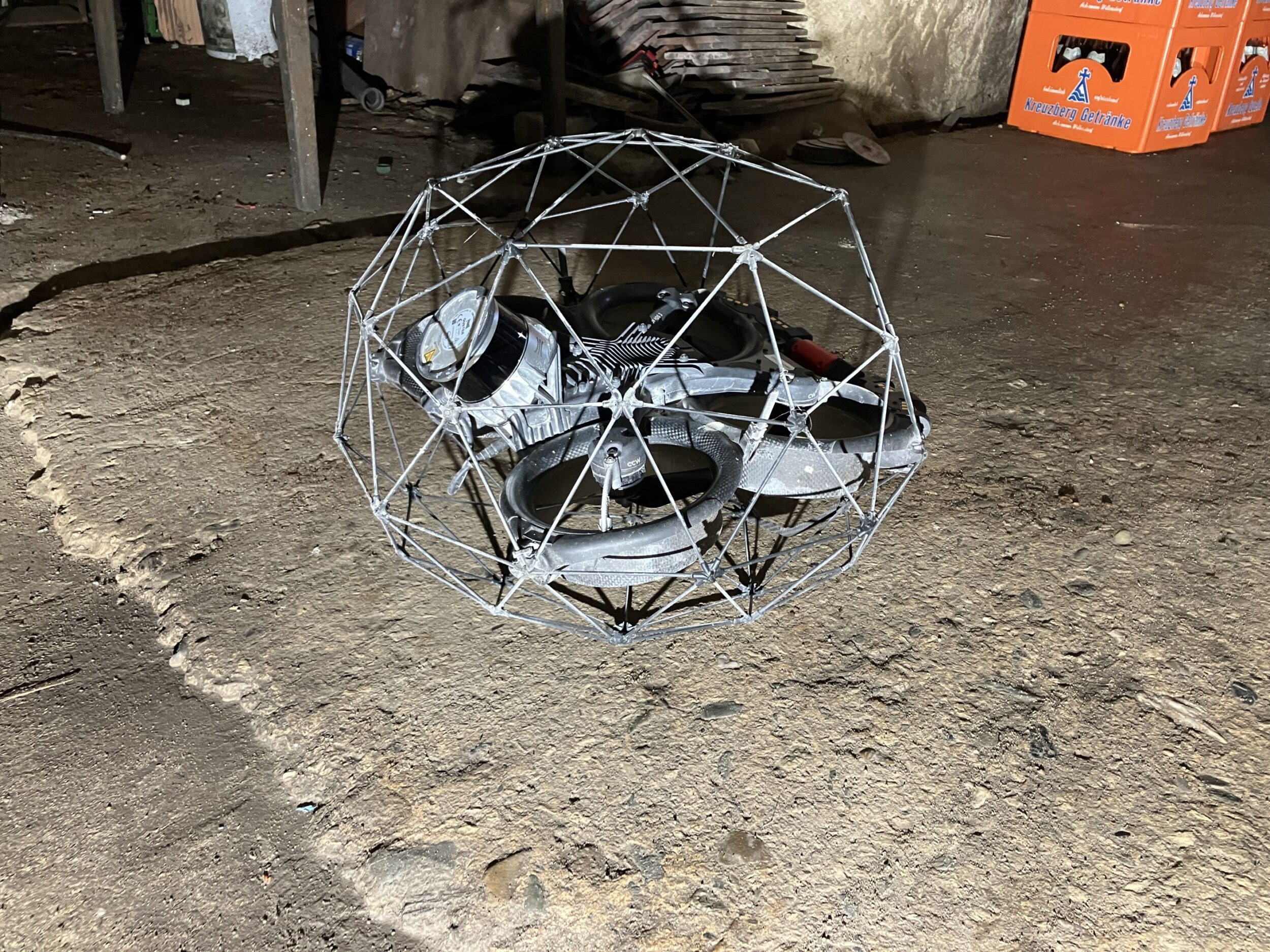 Eine kugelförmig geschützte Drohne liegt auf einem rauen Betonboden in einem Keller. Die Drohne besitzt vier Propeller und ist für den Einsatz in engen oder industriellen Innenräumen konzipiert. Im Hintergrund stehen orangefarbene Getränkekisten und weitere Gegenstände.