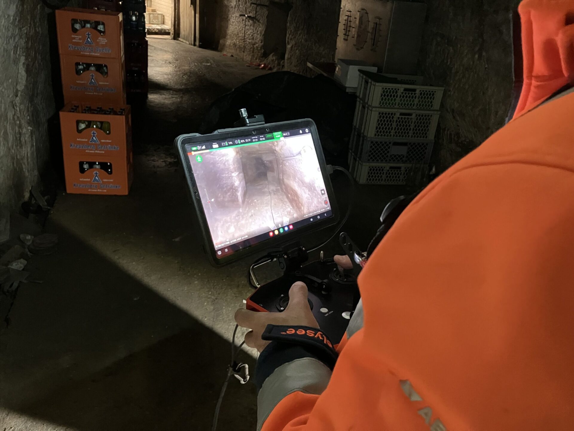 Das Bild zeigt eine Person in einer orangefarbenen Arbeitsjacke, die ein Steuergerät mit großem Bildschirm hält. Auf dem Monitor ist ein Livebild eines Felsenkellers von einer Drohnenkamera zu sehen. Im Hintergrund stehen Getränkekisten und Kunststoffboxen in einem schwach beleuchteten Keller.