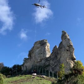 Auf dem Bild fliegt ein Hubschrauber über einer markanten Felsformation mit mehreren steilen Spitzen. Vom Hubschrauber hängt ein langes Seil herab, an dem Lasten befestigt sind, die offenbar für Arbeiten am Felsen genutzt werden. Im Vordergrund ist eine grüne Wiese mit einigen Sträuchern zu sehen, und der Himmel ist klar blau mit wenigen Wolken.