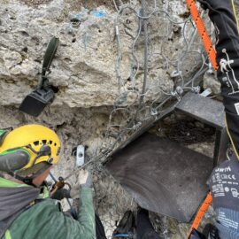 Auf dem Bild ist eine Person mit Schutzhelm und Gehörschutz zu sehen, die an einem steilen Felsen arbeitet. Die Person befestigt eine Vorrichtung, vermutlich eine Wildkamera, an einem Drahtgeflecht, das den Felsen sichert. Daneben ist eine dunkle Plattform montiert, die als Sitz- oder Nistfläche dient, und mehrere Kletterseile sind sichtbar, was auf Arbeiten in großer Höhe hinweist.