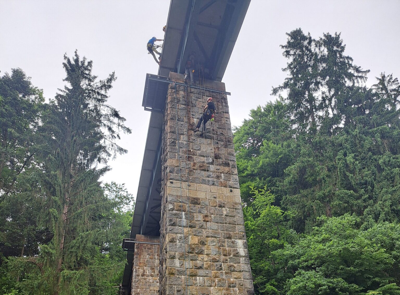 Das Bild zeigt eine hohe Brückenkonstruktion aus Stahl und Stein, die von einem massiven gemauerten Pfeiler getragen wird. An dem Pfeiler hängen mehrere Personen in Schutzkleidung und Helmen, die mit Seilen gesichert sind und offenbar Arbeiten oder ein Training in der Höhe durchführen. Die Umgebung ist von dichtem, grünem Wald mit hohen Bäumen umgeben, was auf eine natürliche, bewaldete Landschaft hinweist.