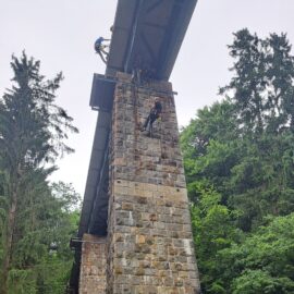Das Bild zeigt eine hohe Brückenkonstruktion aus Stahl und Stein, die von einem massiven gemauerten Pfeiler getragen wird. An dem Pfeiler hängen mehrere Personen in Schutzkleidung und Helmen, die mit Seilen gesichert sind und offenbar Arbeiten oder ein Training in der Höhe durchführen. Die Umgebung ist von dichtem, grünem Wald mit hohen Bäumen umgeben, was auf eine natürliche, bewaldete Landschaft hinweist.