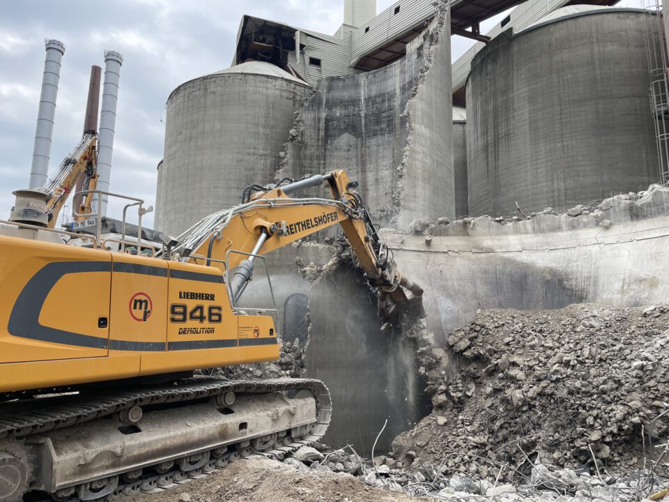 Das Bild zeigt ein großes gelbes Kettenfahrzeug mit einem hydraulischen Greifarm, das an einer massiven Betonstruktur arbeitet. Der Greifarm bricht Teile der Wand ab, wodurch ein Haufen von Schutt und Betonbrocken im Vordergrund liegt. Im Hintergrund sind mehrere große, zylindrische Silos und industrielle Gebäude zu sehen, die auf eine Abriss- oder Rückbauarbeit in einem Kraftwerksbereich hinweisen.