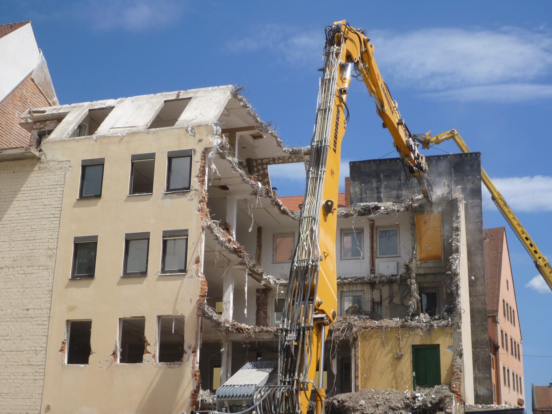 Das Bild zeigt den Abriss eines mehrstöckigen Gebäudes, bei dem ein großer gelber Bagger mit einem hydraulischen Werkzeug die Fassade bearbeitet. Teile der Außenwand sind bereits entfernt, sodass die Innenräume und Etagen sichtbar sind, mit herausragenden Stahlarmierungen und Trümmern. Im Hintergrund sind weitere Häuser mit roten Dächern unter einem blauen Himmel zu sehen.