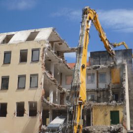 Das Bild zeigt den Abriss eines mehrstöckigen Gebäudes, bei dem ein großer gelber Bagger mit einem hydraulischen Werkzeug die Fassade bearbeitet. Teile der Außenwand sind bereits entfernt, sodass die Innenräume und Etagen sichtbar sind, mit herausragenden Stahlarmierungen und Trümmern. Im Hintergrund sind weitere Häuser mit roten Dächern unter einem blauen Himmel zu sehen.