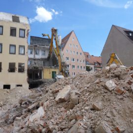 Das Bild zeigt eine große Menge an Bauschutt aus Ziegeln und Beton, die sich als Hügel im Vordergrund auftürmt. Dahinter sind mehrere gelbe Abrissbagger zu sehen, die an einem teilweise abgerissenen Gebäude arbeiten. Im Hintergrund stehen weitere Wohnhäuser mit roten Dächern und hellen Fassaden unter einem blauen Himmel mit wenigen Wolken.