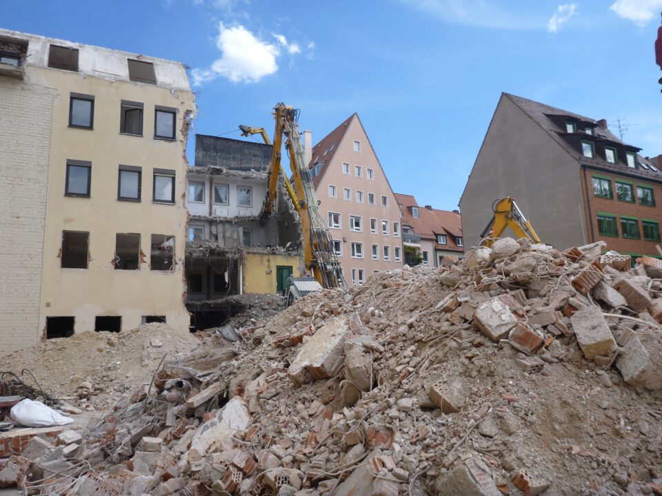 Das Bild zeigt eine große Menge an Bauschutt aus Ziegeln und Beton, die sich als Hügel im Vordergrund auftürmt. Dahinter sind mehrere gelbe Abrissbagger zu sehen, die an einem teilweise abgerissenen Gebäude arbeiten. Im Hintergrund stehen weitere Wohnhäuser mit roten Dächern und hellen Fassaden unter einem blauen Himmel mit wenigen Wolken.