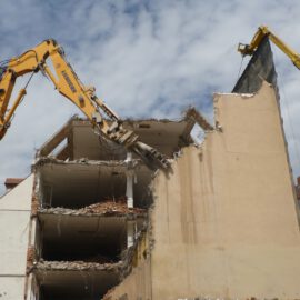 Das Bild zeigt den Abriss eines mehrstöckigen Gebäudes, bei dem zwei große gelbe Bagger mit langen Auslegern im Einsatz sind. Einer der Bagger greift mit einem hydraulischen Werkzeug die obere Ecke der Fassade, während der andere eine Schutzwand hält. Die freigelegte Gebäudestruktur zeigt offene Etagen mit Trümmern und herausragenden Stahlteilen, im Hintergrund sind weitere Häuser mit roten Dächern zu sehen.