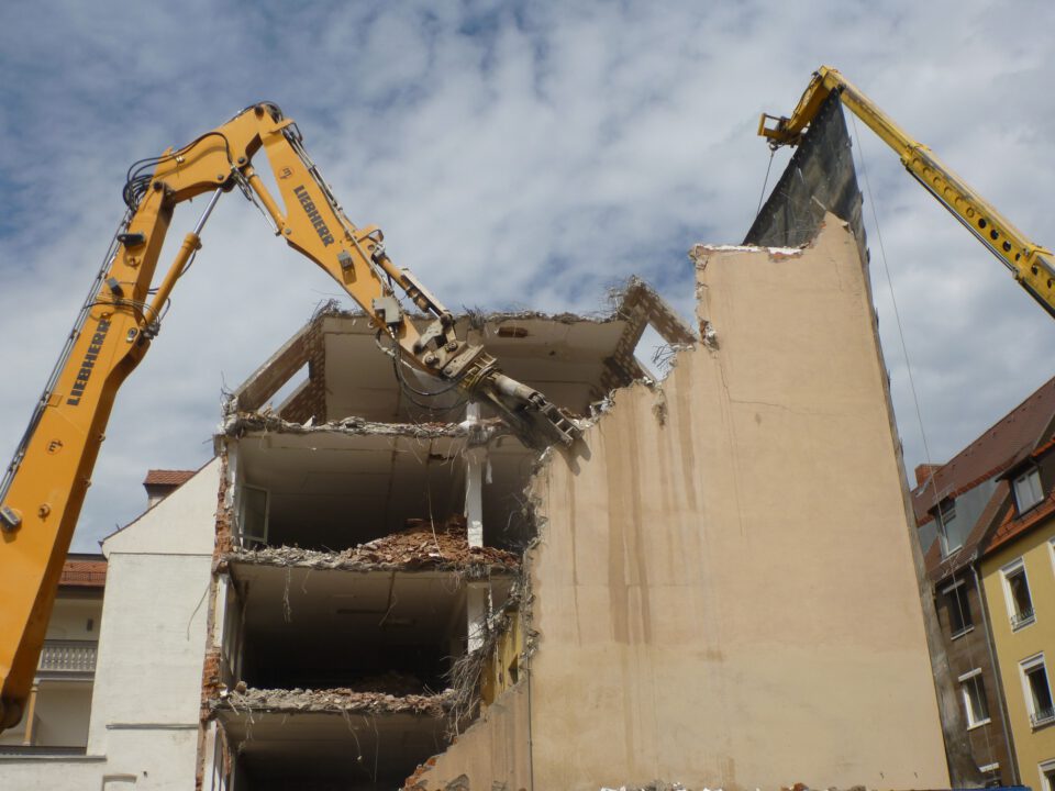 Das Bild zeigt den Abriss eines mehrstöckigen Gebäudes, bei dem zwei große gelbe Bagger mit langen Auslegern im Einsatz sind. Einer der Bagger greift mit einem hydraulischen Werkzeug die obere Ecke der Fassade, während der andere eine Schutzwand hält. Die freigelegte Gebäudestruktur zeigt offene Etagen mit Trümmern und herausragenden Stahlteilen, im Hintergrund sind weitere Häuser mit roten Dächern zu sehen.
