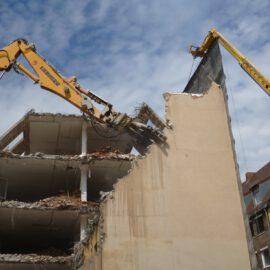 Das Bild zeigt den Abriss eines mehrstöckigen Gebäudes, bei dem zwei große gelbe Bagger mit langen Auslegern im Einsatz sind. Einer der Bagger greift mit einem hydraulischen Werkzeug die obere Ecke der Fassade, während der andere eine Schutzwand hält. Die freigelegte Gebäudestruktur zeigt offene Etagen mit Trümmern und herausragenden Stahlteilen, im Hintergrund sind weitere Häuser mit roten Dächern zu sehen.