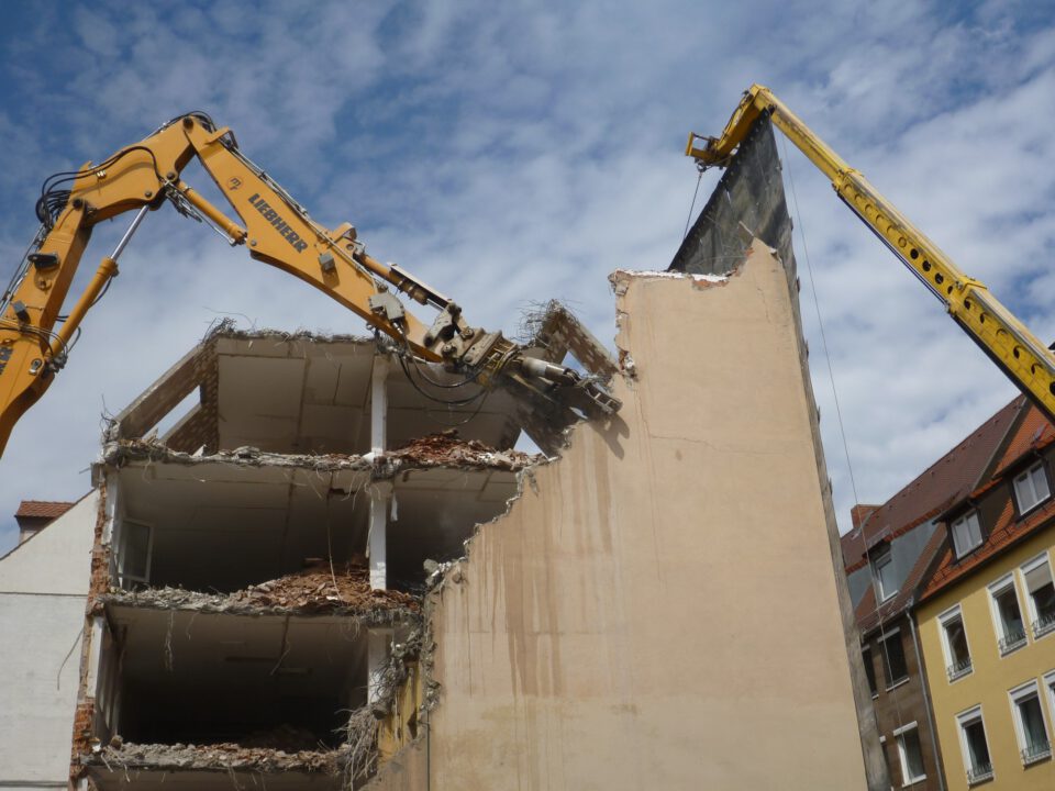 Das Bild zeigt den Abriss eines mehrstöckigen Gebäudes, bei dem zwei große gelbe Bagger mit langen Auslegern im Einsatz sind. Einer der Bagger greift mit einem hydraulischen Werkzeug die obere Ecke der Fassade, während der andere eine Schutzwand hält. Die freigelegte Gebäudestruktur zeigt offene Etagen mit Trümmern und herausragenden Stahlteilen, im Hintergrund sind weitere Häuser mit roten Dächern zu sehen.