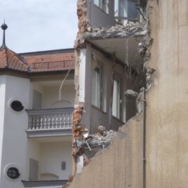 Das Bild zeigt den Teilabriss eines mehrstöckigen Gebäudes, bei dem die rechte Hälfte der Fassade fehlt und die Innenräume sichtbar sind. Die freigelegte Wand zeigt zerbrochene Betonplatten, herausragende Stahlarmierungen und lose Trümmer. Im Hintergrund ist ein intaktes Gebäude mit hellem Putz, runden Fenstern und einem roten Ziegeldach zu sehen.