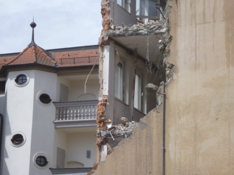 Das Bild zeigt den Teilabriss eines mehrstöckigen Gebäudes, bei dem die rechte Hälfte der Fassade fehlt und die Innenräume sichtbar sind. Die freigelegte Wand zeigt zerbrochene Betonplatten, herausragende Stahlarmierungen und lose Trümmer. Im Hintergrund ist ein intaktes Gebäude mit hellem Putz, runden Fenstern und einem roten Ziegeldach zu sehen.