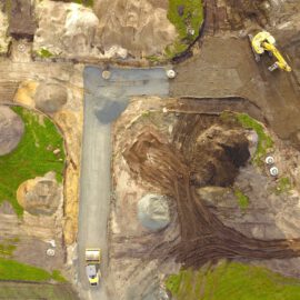 Das Bild zeigt eine Baustelle aus der Vogelperspektive mit mehreren Bereichen, die für Erdarbeiten vorbereitet sind. Im oberen Teil ist ein gelber Bagger zu sehen, der auf einer dunklen Erdfläche arbeitet, während im unteren Bereich ein Lastwagen auf einer Schotterstraße steht. Rund um die Baustelle liegen große Erdhaufen und grüne Grasflächen, und verschiedene runde Betonabdeckungen sind im Boden verteilt.