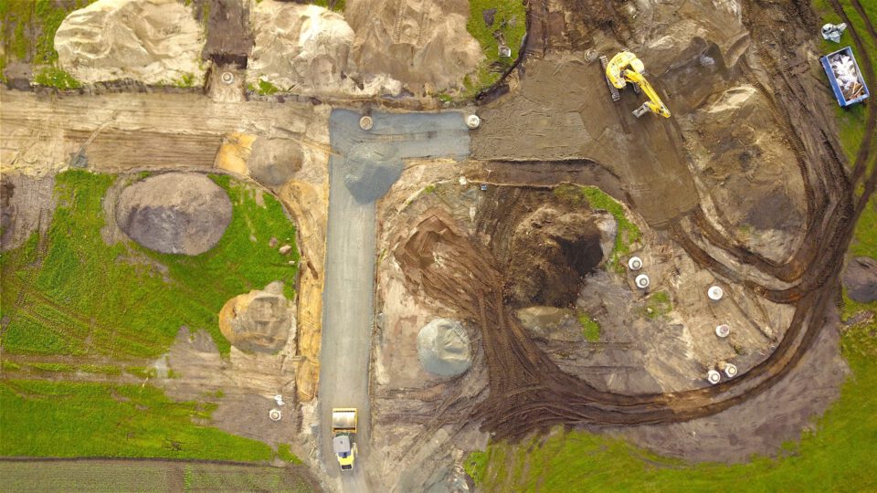 Das Bild zeigt eine Baustelle aus der Vogelperspektive mit mehreren Bereichen, die für Erdarbeiten vorbereitet sind. Im oberen Teil ist ein gelber Bagger zu sehen, der auf einer dunklen Erdfläche arbeitet, während im unteren Bereich ein Lastwagen auf einer Schotterstraße steht. Rund um die Baustelle liegen große Erdhaufen und grüne Grasflächen, und verschiedene runde Betonabdeckungen sind im Boden verteilt.