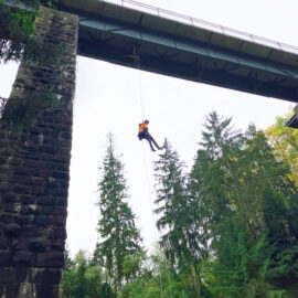 Das Bild zeigt eine Person, die an einem Seil von einer hohen Brücke abseilt. Die Brücke besteht aus einer massiven Stahlkonstruktion mit steinernen Pfeilern, die sich zwischen hohen Nadelbäumen erheben. Im Hintergrund ist dichter Wald zu sehen, und die Szene spielt bei Tageslicht unter einem leicht bewölkten Himmel.