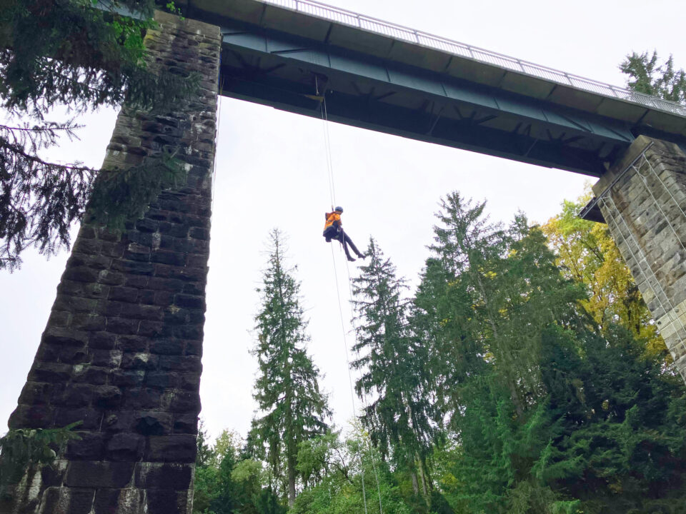 Das Bild zeigt eine Person, die an einem Seil von einer hohen Brücke abseilt. Die Brücke besteht aus einer massiven Stahlkonstruktion mit steinernen Pfeilern, die sich zwischen hohen Nadelbäumen erheben. Im Hintergrund ist dichter Wald zu sehen, und die Szene spielt bei Tageslicht unter einem leicht bewölkten Himmel.