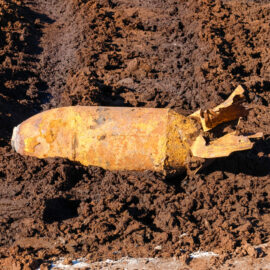 Das Bild zeigt eine stark verrostete Fliegerbombe, die teilweise aus brauner Erde freigelegt wurde. Die Bombe hat eine zylindrische Form mit einer spitzen Vorderseite und einem beschädigten Leitwerk am hinteren Ende. Die Oberfläche ist unregelmäßig und von Rost und Erdanhaftungen bedeckt, was auf ein langes Verweilen im Boden hinweist.
