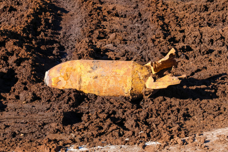 Das Bild zeigt eine stark verrostete Fliegerbombe, die teilweise aus brauner Erde freigelegt wurde. Die Bombe hat eine zylindrische Form mit einer spitzen Vorderseite und einem beschädigten Leitwerk am hinteren Ende. Die Oberfläche ist unregelmäßig und von Rost und Erdanhaftungen bedeckt, was auf ein langes Verweilen im Boden hinweist.