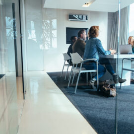 Das Bild zeigt einen modernen Besprechungsraum mit Glaswänden, in dem mehrere Personen an einem Tisch sitzen. Auf dem Tisch steht ein Laptop, und die Gruppe scheint sich in einem Gespräch oder einer Besprechung zu befinden. Der Raum ist hell und minimalistisch eingerichtet, mit großen Fenstern, die Tageslicht hereinlassen, und einem Teppichboden, der den Bereich gemütlich wirken lässt.