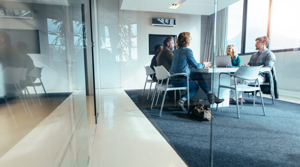 Das Bild zeigt einen modernen Besprechungsraum mit Glaswänden, in dem mehrere Personen an einem Tisch sitzen. Auf dem Tisch steht ein Laptop, und die Gruppe scheint sich in einem Gespräch oder einer Besprechung zu befinden. Der Raum ist hell und minimalistisch eingerichtet, mit großen Fenstern, die Tageslicht hereinlassen, und einem Teppichboden, der den Bereich gemütlich wirken lässt.
