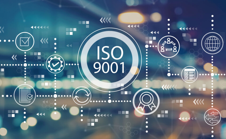 Das Bild zeigt eine grafische Darstellung mit dem Schriftzug ISO 9001 in einem großen Kreis in der Mitte. Rund um diesen Kreis sind verschiedene Symbole angeordnet, die Themen wie Qualitätssicherung, Dokumentation, Prozesse und Zertifizierung darstellen. Der Hintergrund ist unscharf und zeigt helle Lichtpunkte auf einem dunklen, bläulichen Untergrund, was der Darstellung einen modernen und technologischen Charakter verleiht.