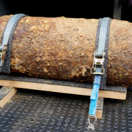 Das Bild zeigt eine große, stark verrostete Bombe, die auf einer Metallfläche liegt. Sie ist mit zwei blauen Spanngurten auf einem Holzgestell fixiert, um sicheren Halt zu gewährleisten. Die Oberfläche der Bombe ist uneben und von Rost und Ablagerungen bedeckt, was auf ein hohes Alter hinweist.