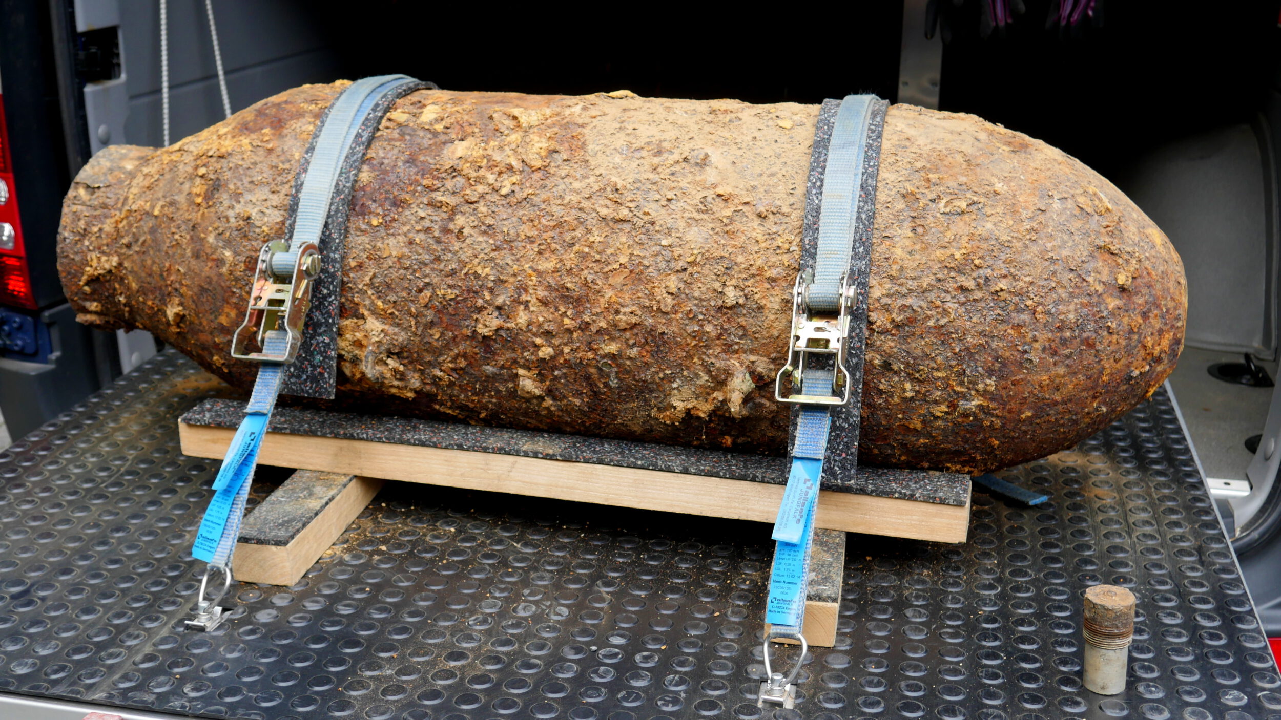 Das Bild zeigt eine große, stark verrostete Bombe, die auf einer Metallfläche liegt. Sie ist mit zwei blauen Spanngurten auf einem Holzgestell fixiert, um sicheren Halt zu gewährleisten. Die Oberfläche der Bombe ist uneben und von Rost und Ablagerungen bedeckt, was auf ein hohes Alter hinweist.