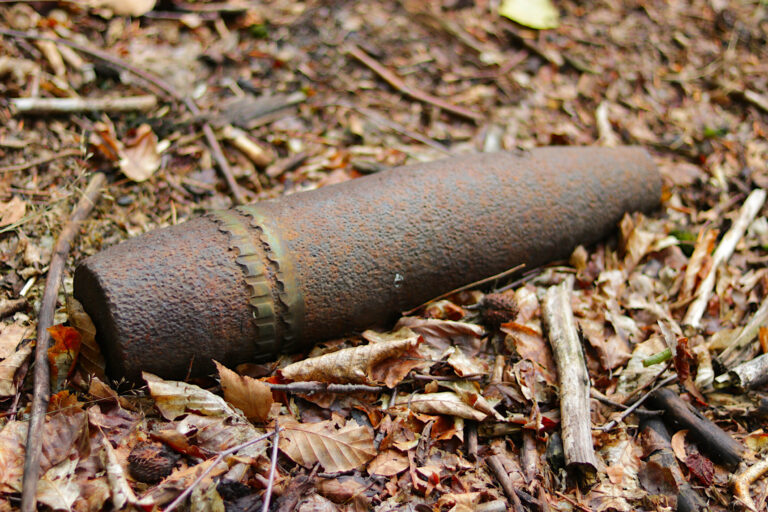 Das Bild zeigt eine stark verrostete, längliche Metallhülle, die auf dem Waldboden liegt. Sie ist von trockenen Blättern und kleinen Ästen umgeben, was auf eine natürliche Umgebung hinweist. Die Form und Struktur lassen erkennen, dass es sich um ein altes, vermutlich militärisches Geschoss handelt.